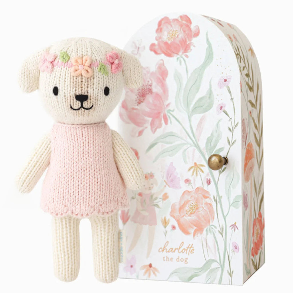 A knitted teddy bear dog with a pink dress and flower crown stands in front of its floral-patterned gift box. The box features pink flowers, green leaves, and the text 'charlotte the dog' with a small gold clasp.