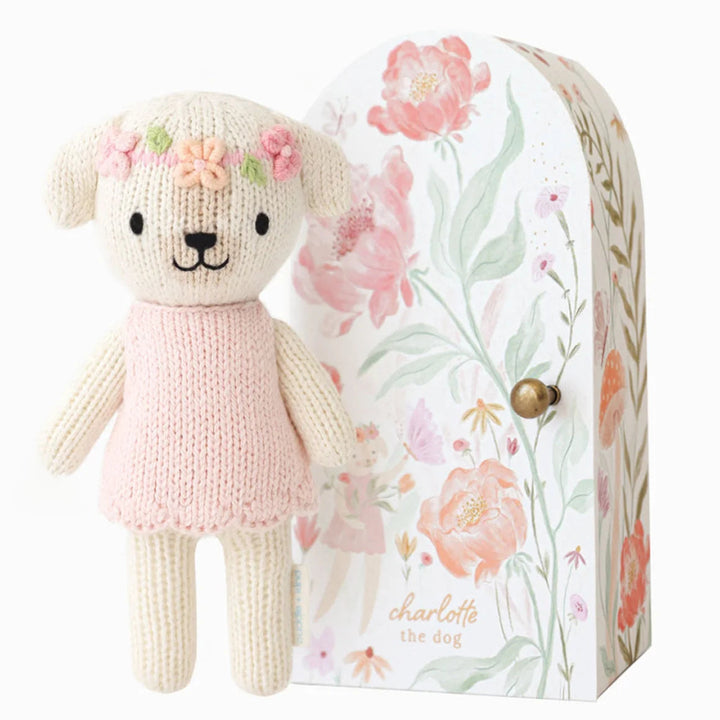 A knitted teddy bear dog with a pink dress and flower crown stands in front of its floral-patterned gift box. The box features pink flowers, green leaves, and the text 'charlotte the dog' with a small gold clasp.