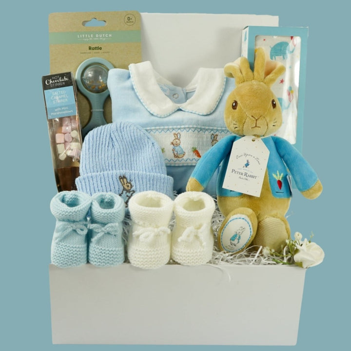 white baby hamper box with Beatrix potter  Peter rabbit soft toy, blue velour baby sleepsuit with smocked yolk with bunny's , blue beanie hat with bunny,  Little Dutch baby rattle, white muslin swaddle with blue elephants, salted caramel chocolate spoon with marshmallow