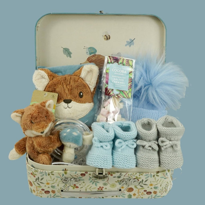 cardboard suitcase with soft activity book in blue with fox head design, fox soft rattle, chocolate spoon with mallows, blue fluffy pompom hat, two pairs of booties 