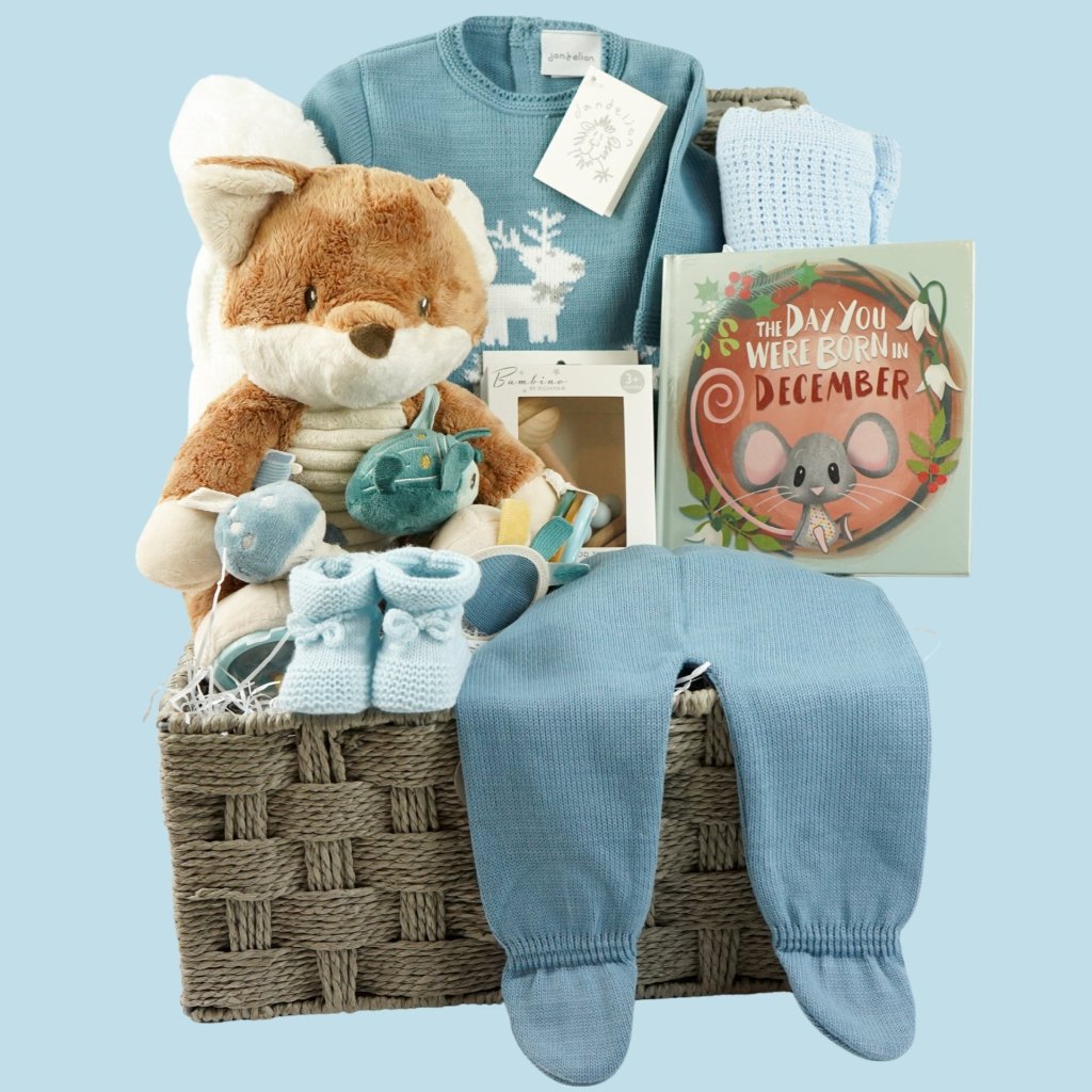 baby boy Christmas hamper gifts in a grey hamper basket includes blue knit outfit with white reindeer and snowflake design, blue cellular blanket, wooden rattle , blue booties , white bobble hat, cellular blanket, Born in December book