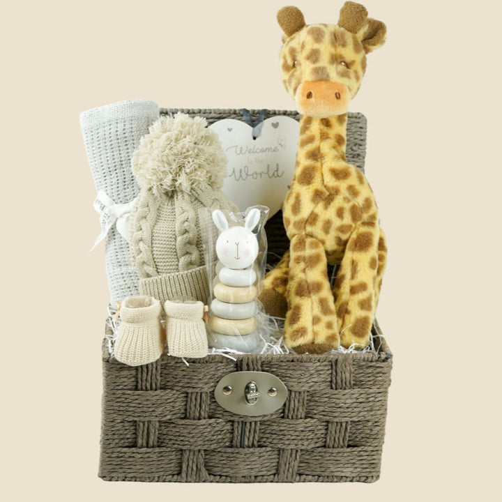 Grey hamper basket with baby gifts including grey cellular blanket, biscuit coloured knit pompom hat, wooden stacking toy, nursery plaque in heart shape with 'welcome to the world', soft giraffe