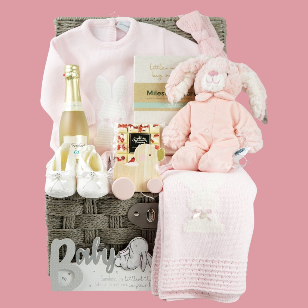 baby girl hamper basket includes fine knit pink romper with feet and white bunny  and matching blanket, small bottle alcohol free sparkling wine, satin baby shoes, white chocolate eton mess bar, wooden push along duck, pink bunny in velour romper, memorable moments cars in a box, baby plaque