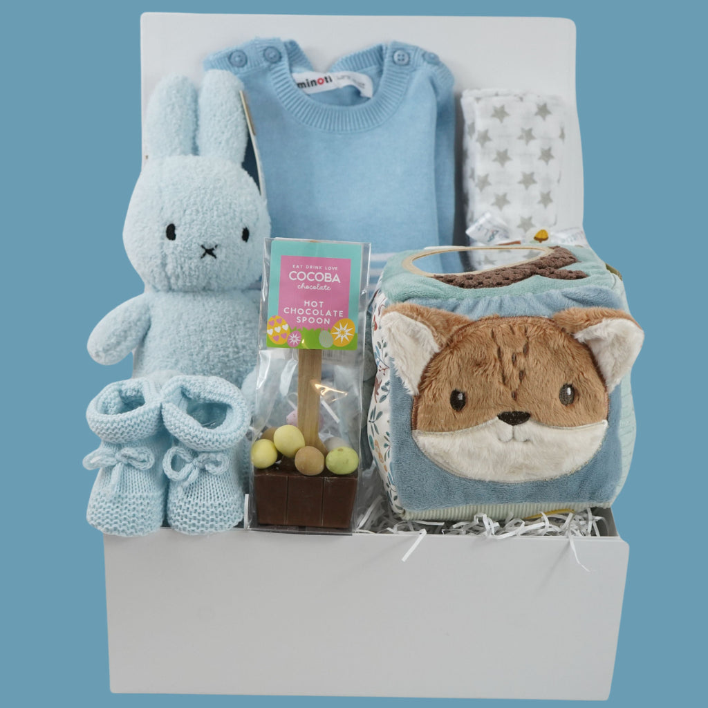 baby boy hamper with blue fine knit romper with white striped and pocket, white muslin with silver stars, blue terry miffy, Forest friends Fox soft activity book, blue knit booties, chocolate stirrer spoon with mini eggs 