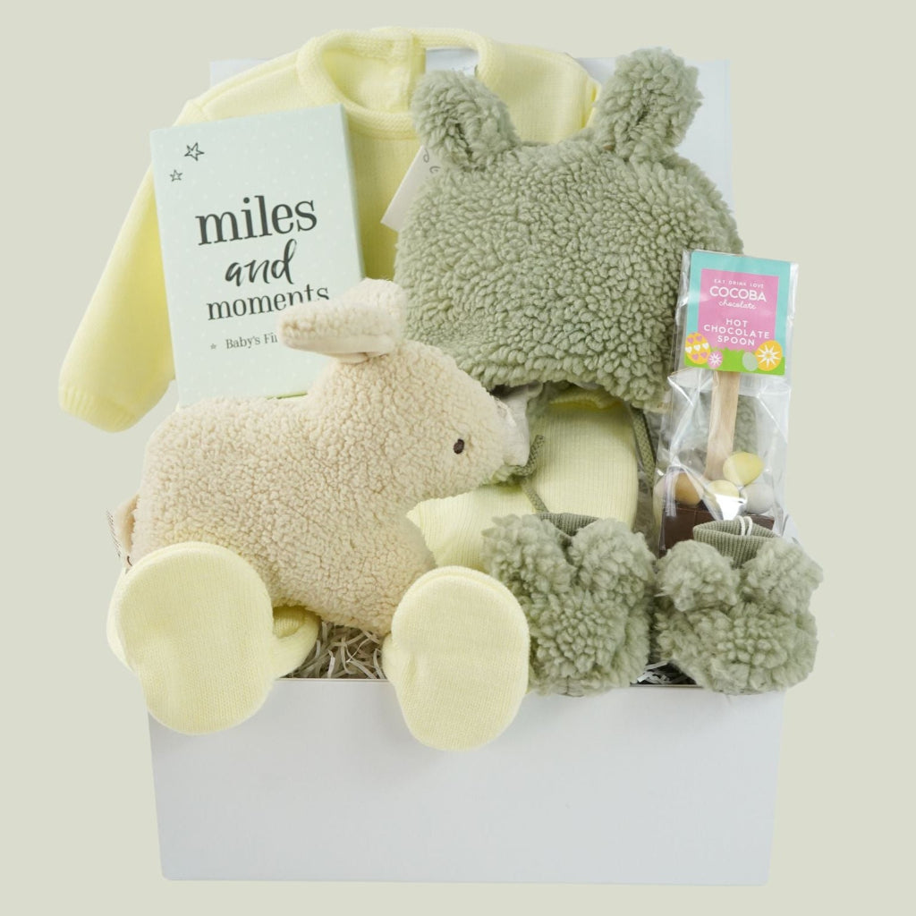 easter baby hamper, yellow knit romper with bunny design and pompom tail, soft fluffy bunny ears green hat with matching bunny ears , booties , recycled cream bunny cuddle toy, miles and moments baby cards, hot chocolate spoon with small eggs 