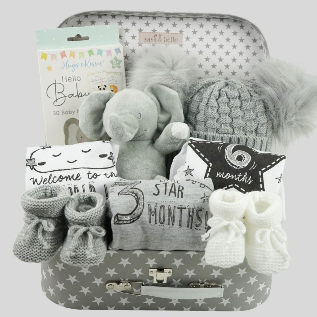 grey storage suitcase with baby items including baby hat with soft fluffy double pompoms, baby memory cards, soft elephant toy, baby set of milestone vests , white knit booties and grey knit booties