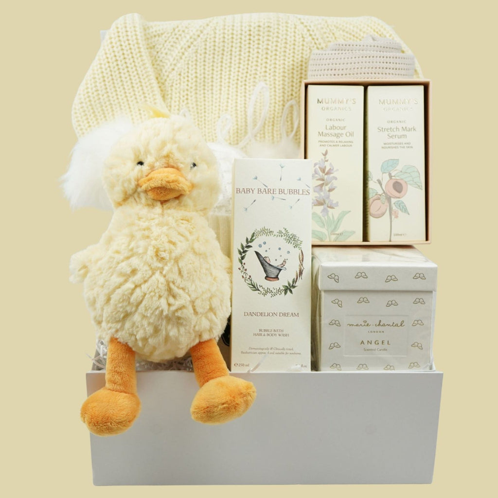 A baby gift basket set containing a fluffy cream plush duck toy, a cream knit blanket, and several boxed baby items including a 'Baby Gear in Bed' book and 'Angel' chocolates, arranged in a white basket against a solid pale yellow background.
