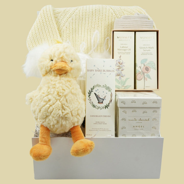 A baby gift basket set containing a fluffy cream plush duck toy, a cream knit blanket, and several boxed baby items including a 'Baby Gear in Bed' book and 'Angel' chocolates, arranged in a white basket against a solid pale yellow background.