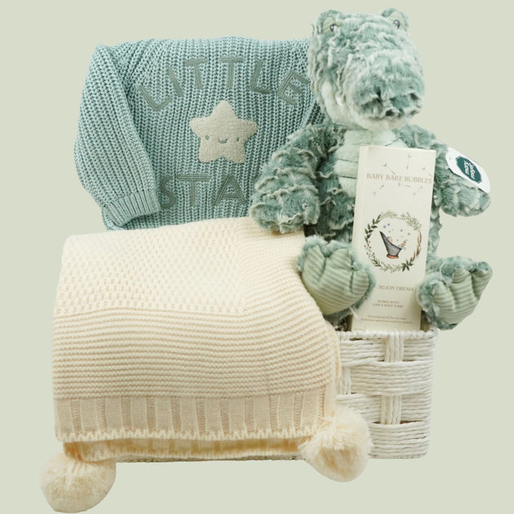 A baby gift set featuring a light blue knit sweater with a star patch, a cream knit blanket with pom-poms, and a plush green teddy bear holding a tag, arranged in a white basket against a solid light green background.