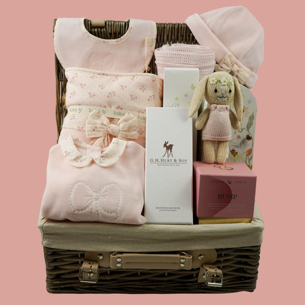 A baby gift basket arranged in a wicker suitcase with a light beige fabric lining, filled with soft pink baby clothing, folded textiles, a small plush bunny, and gift boxes, set against a solid pink background.
