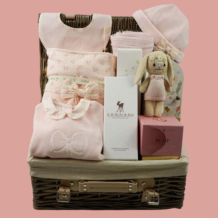 A baby gift basket arranged in a wicker suitcase with a light beige fabric lining, filled with soft pink baby clothing, folded textiles, a small plush bunny, and gift boxes, set against a solid pink background.