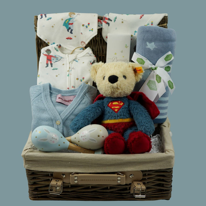 A baby gift basket set containing a blue knit cardigan, a plush teddy bear dressed as Superman, a blue blanket with a white star, patterned baby socks, and folded baby clothes inside a wicker basket.
