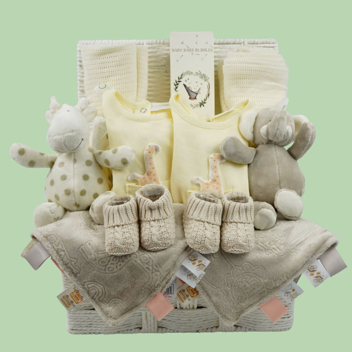 A baby gift basket set containing pale yellow baby clothes, beige knit booties, a plush giraffe toy, a plush elephant toy, and textured beige blankets, arranged in a wicker basket against a solid light green background.