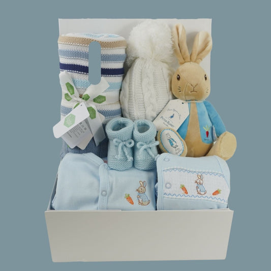 baby hamper with peter rabbit blue outfit including a pale blue sleepsuit and matching jacket, pale blue booties, blue and white and beige baby blanket, white pom pom hat and peter rabbit soft toy