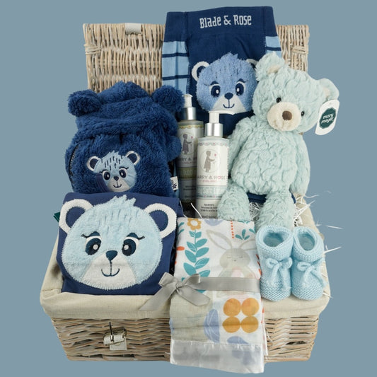 white wicker hamper basket, blue with teddy design baby clothing set includes baby leggings with teddy on the bum, feece jacket with hood in navy with teddy and blue long sleeved top with teddy face, organic toiletries, pale blue teddy, swaddle with teddy and owl design, pale blue knit booties 