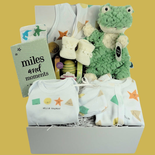 white hamper box with baby gifts including a white cotton layette set with shapes designs on the sleepsuit and vest, baby milestone cards in a green box, white hot chocolate bomb , white muslin with green and blue dinosaurs, white knit baby booties , green frog lovey