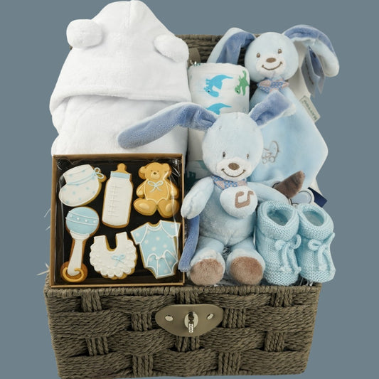 Baby boy hamper basket, white baby dressing gown with ears, muslin with dinosaurs in green and blue, blue Nattou rabbit musicaql and matching comforter, baby booties in blue, box of baby themed biscuits in blue and white