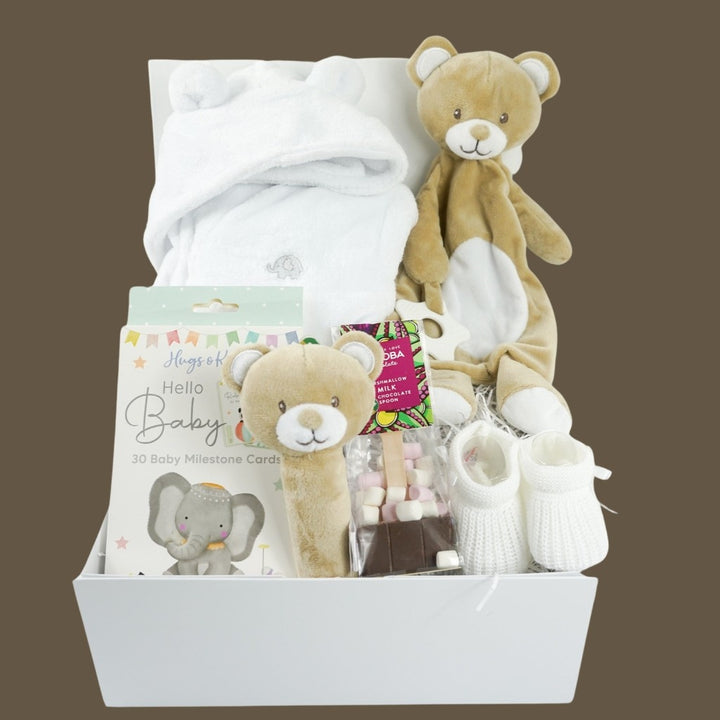 white hamper box with baby items including baby dressing gown in white with cute ears, soft brown baby comforter and matching squeaky rattle, baby milestone cards with cute animals, white knit baby booties , hot chocolate spoon