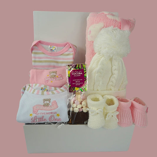 baby girl hamper box with 3 baby vesys with cute designs and pink trim, hot chocolate spoon with marshmallows, baby white cable pom pom hat, white knit booties, pink knit booties , pink baby blanket with white teddy faces