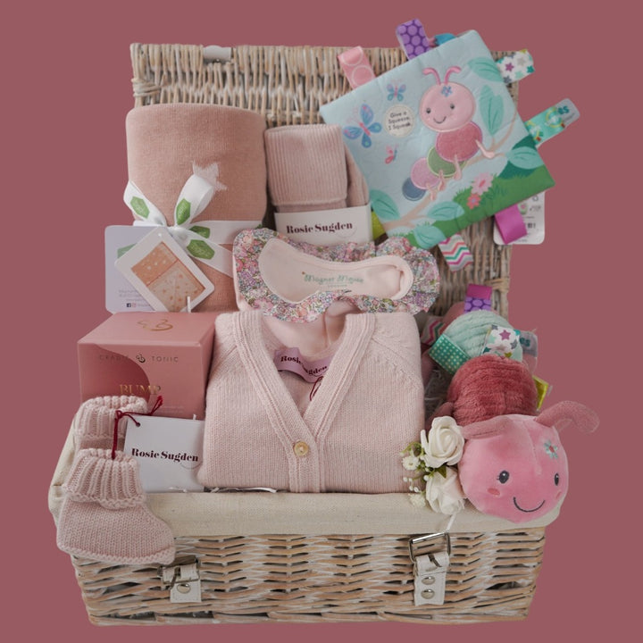 White wash hamper basket, baby gifts including baby pink cashmere cardigan, pink reversible baby blanket with stars, taggie caterpillar soft toy, taggie caterpillar crickle teether book, mum cashmere bed socks in poink, pink bump candle in a gift box, pink cashmere baby booties , pink magnetic closing baby sleepsuit with frilled collar