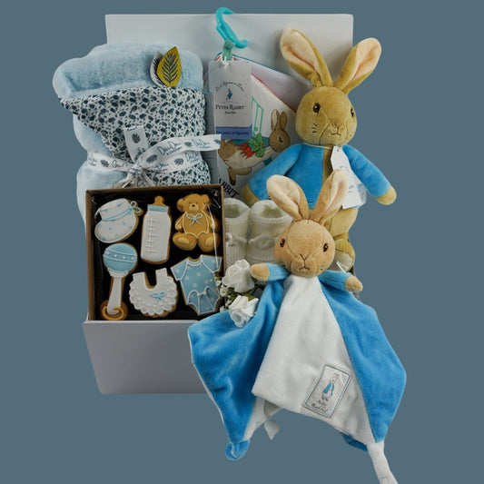 Baby hamper box includes peter rabbit soft toy, Peter rabbit blue and white baby comforter, white baby knit booties, Peter rabbit cloth soft baby book, soft pale blue doudou baby blanket, baby themed celebration biscuits