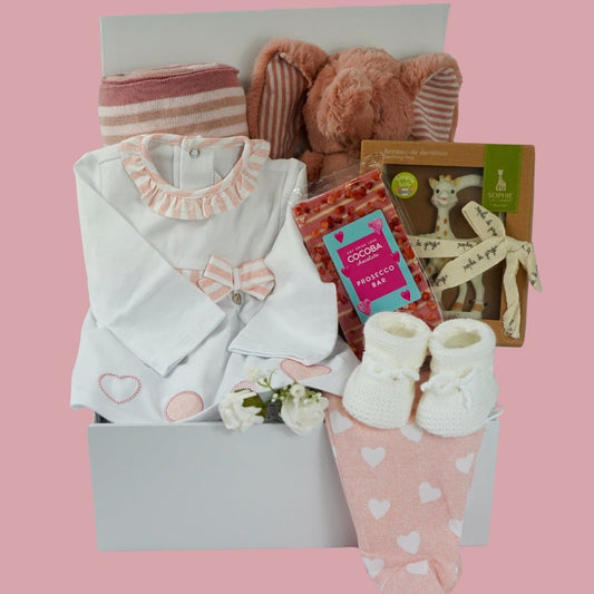 Baby hamper box with pink baby blanket with stars, soft plus elephant in pink and white, baby designer  clothing white top with pink ruffled neck and pink and white bow , pin leggings with white hearts , cocoba prosecco chocolate bar, sophie ;la girafe teether, white baby  booties 