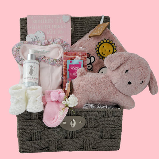 Grey baby hamper box with pink magnetic fastening baby sleepsuit with frilled liberty collar, baby booties, pink snuffie miffy and friends soft dog, soft pink fluffy book, prosecco chocolate bar, organic baby lotion, baby socks in pink with a slogan on the foot and pom poms , white knit booties 