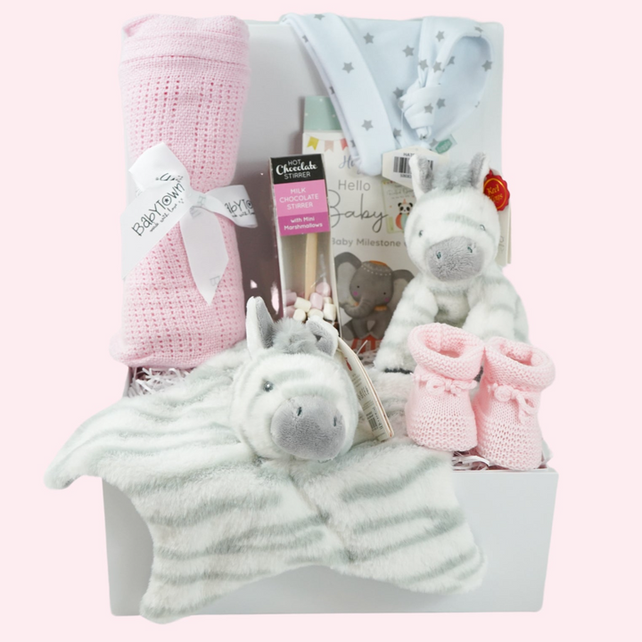 White hamper box with pink cellular baby blanket, white knot hat with silver stars, baby milestone cards, chocolate stirrer spoon with marshmallows, pink knit baby booties , grey and white zebra soft toy and matching comforter