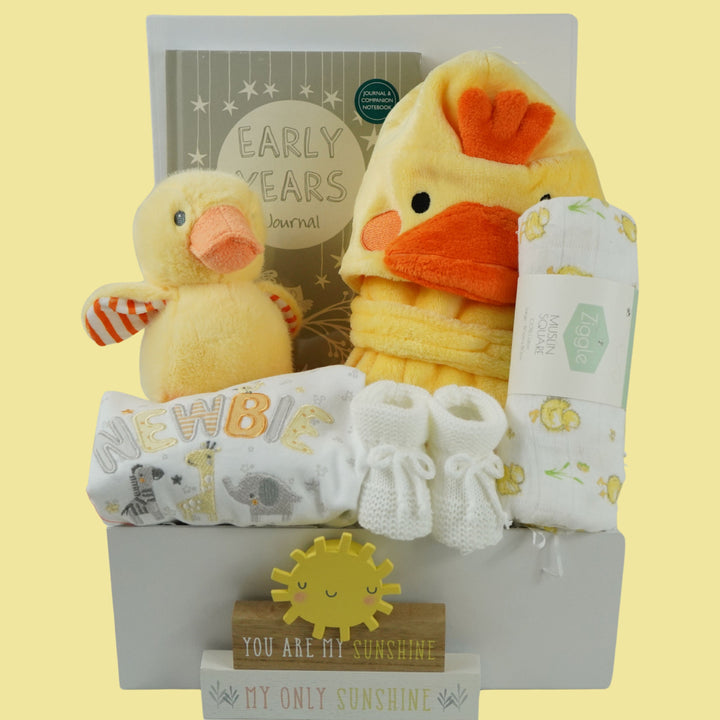 white hamper box with yellow baby dressing gown with duck face hood, soft cuddly yellow duck toy, white muslin square with yellow duck pattern., white knit baby booties, white baby sleep suit with zebra, giraffe and elephant and applique NEWBIE all in grey, yellow and orange, Early years baby journal 