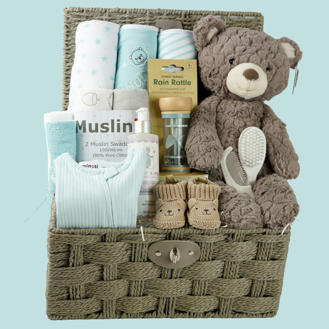 grey paper rope hamper basket with three blue and white hooded towels, baby rain rattle, 2 muslins, 3 wash clothes 
organic baby bath, blue ribbed zip up baby sleepsuit, caramel bunny knit baby shoes, baby brush and comb, soft brown/grey baby teddy
