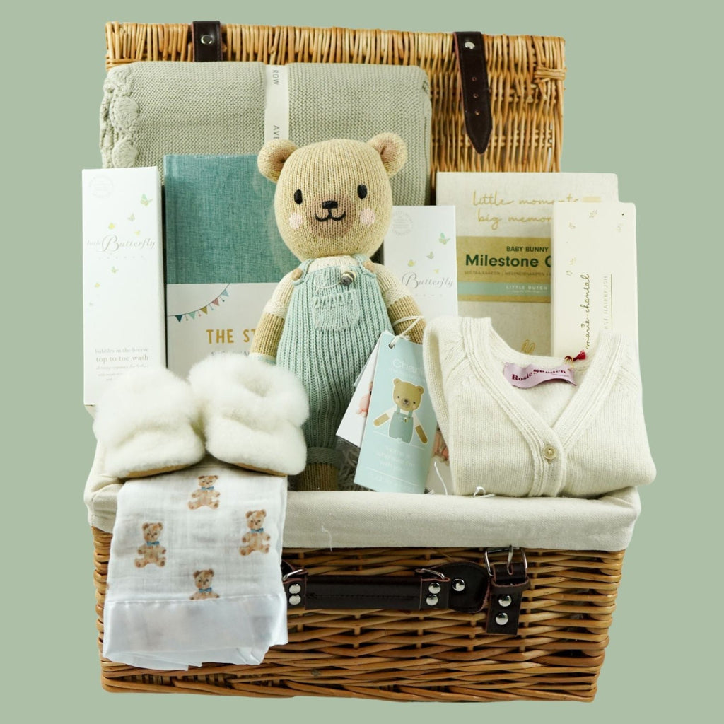 luxury baby boy hamper in a wicker basket with luxury green baby blanket with scalloped edge , baby journal with green cover, soft alpaca baby slippers, Cuddle & Kind soft knit teddy in green dungarees, cashmere baby cardigan and mummy matching cashmere socks, milestone cards and baby toiletries 
