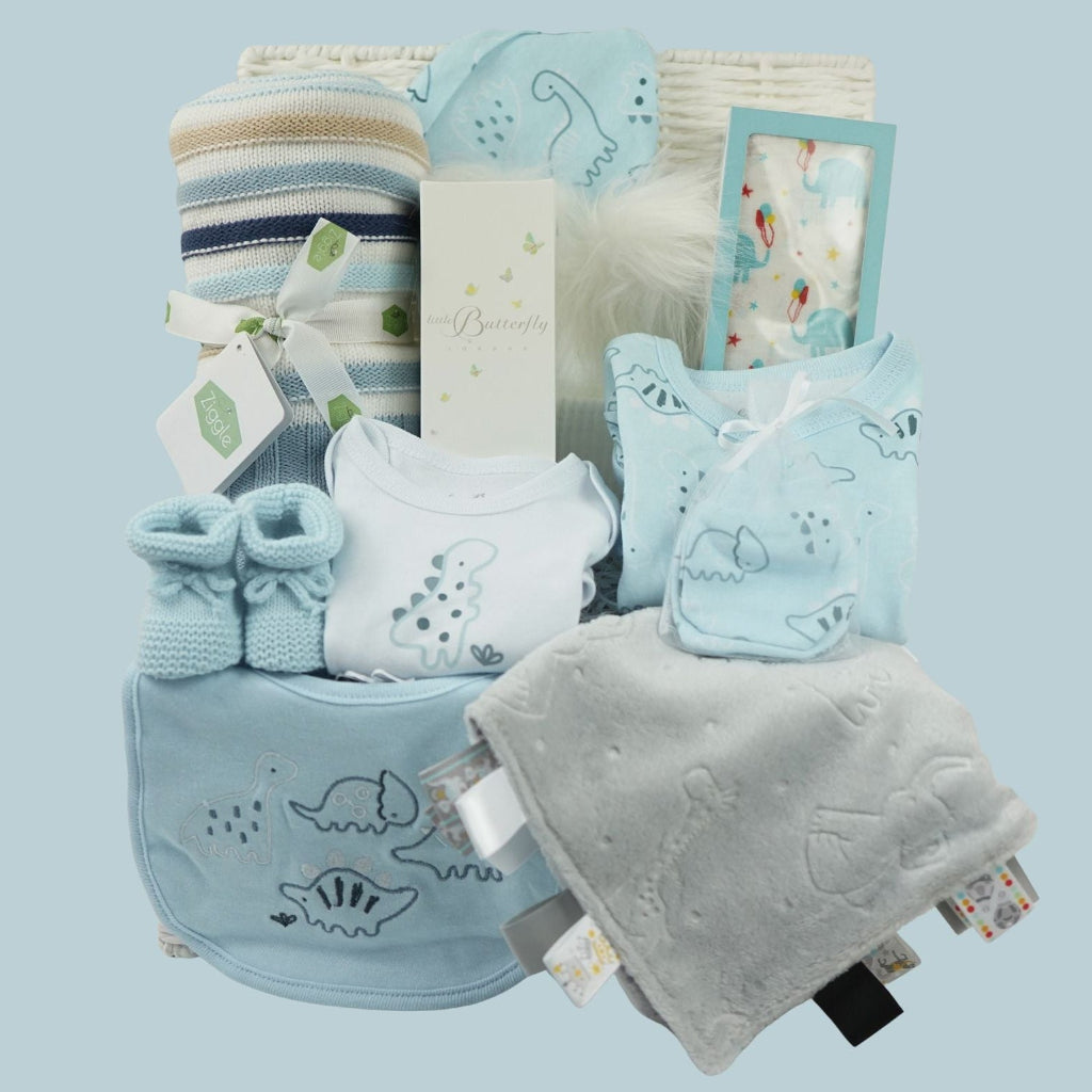 baby boy gift hamper with blue, white and cream shaped blanket, baby dinosaur clothing set, organic baby bubble bath, soft grey taggie comforter, blue booties and white double pom pom baby hat