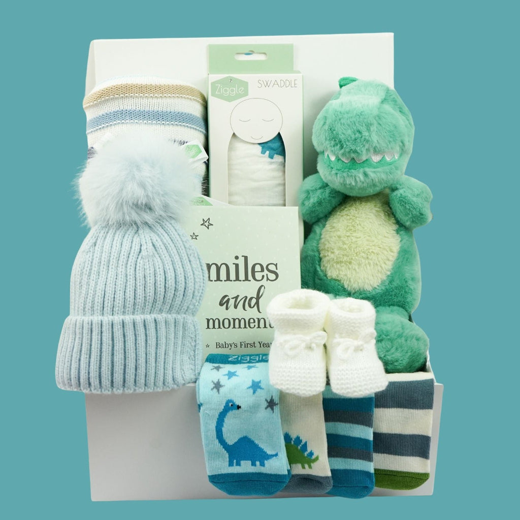 A baby gift set in a white box featuring a light blue knit beanie with a fluffy pom-pom, a green plush dinosaur toy, a pair of white booties, and several patterned socks in blue, white, and navy.
