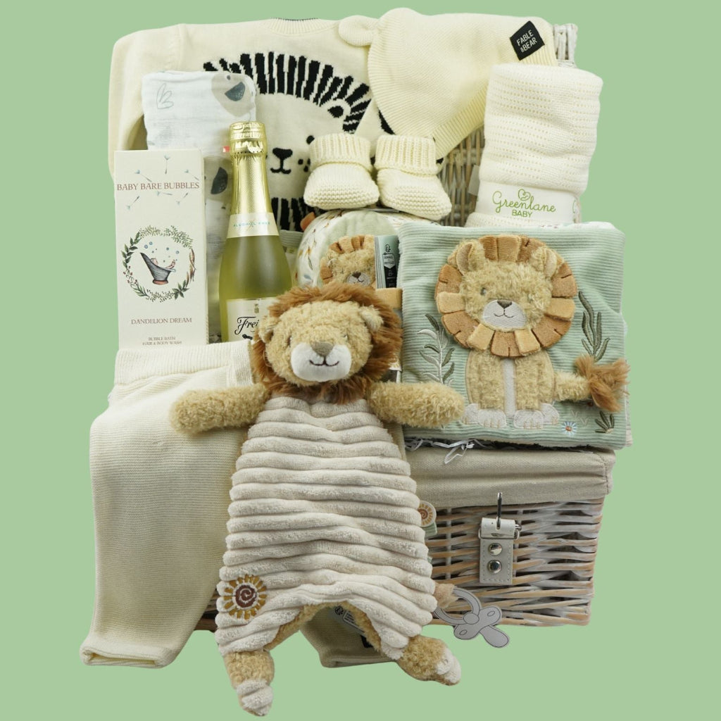 A baby gift hamper set against a solid sage green background, containing a wicker basket filled with baby items including a plush lion lovey, folded cream and mint green blankets, a small bottle, and a champagne flute.