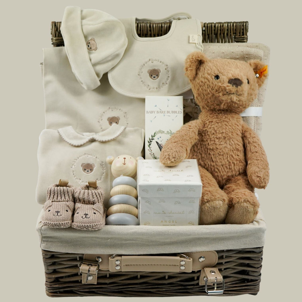 A baby gift basket set containing a brown teddy bear, a cream bib, a cream sleepsuit with embroidered teddy and matching baby wrap, a pair of cream booties, pastel stacking toy, and a small gift box, arranged inside a wicker basket 
