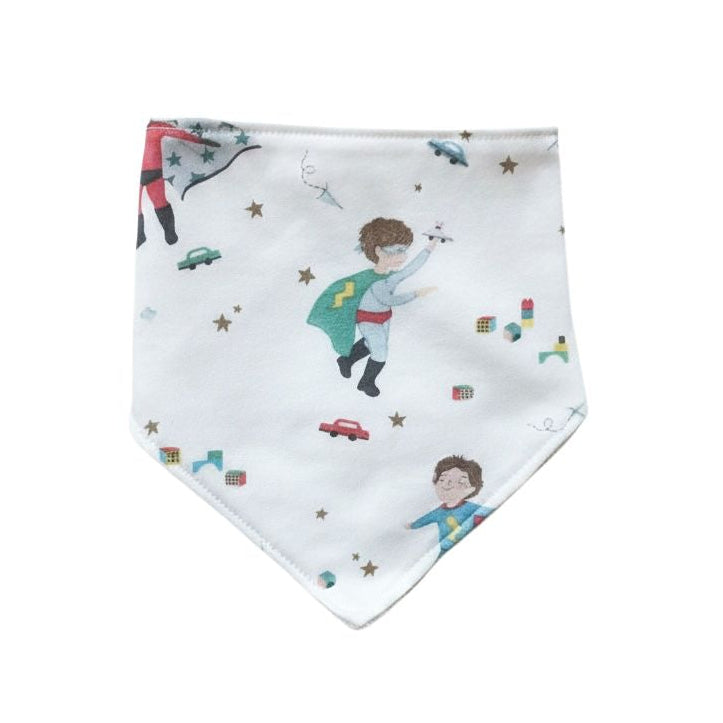 A white baby bandana bib featuring a whimsical print of children in superhero costumes, stars, and small toy cars, displayed flat against a solid white background.