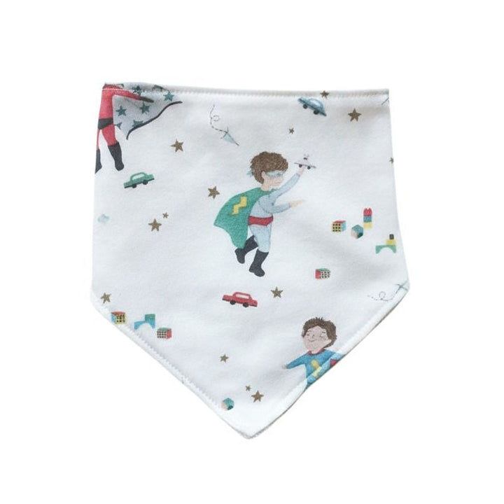 A white baby bandana bib featuring a whimsical print of children in superhero costumes, stars, and small toy cars, displayed flat against a solid white background.