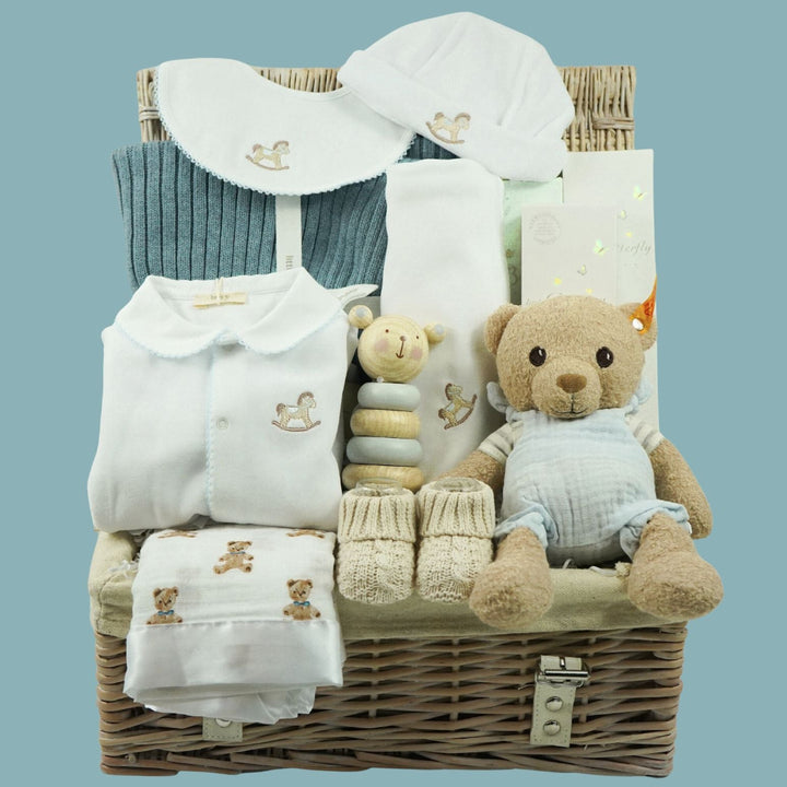 A baby gift basket set containing a blue knit baby sweater, white baby clothes with embroidered details, a plush teddy bear, a small stuffed animal, a stack of wooden teethers, and a hat, all arranged inside a wicker basket against a solid blue background.