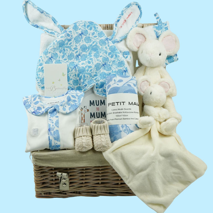A baby gift basket containing a blue and white patterned bonnet with bunny ears, a white plush mouse lovey, a white muslin swaddle blanket, a pair of beige booties, and a wicker basket.