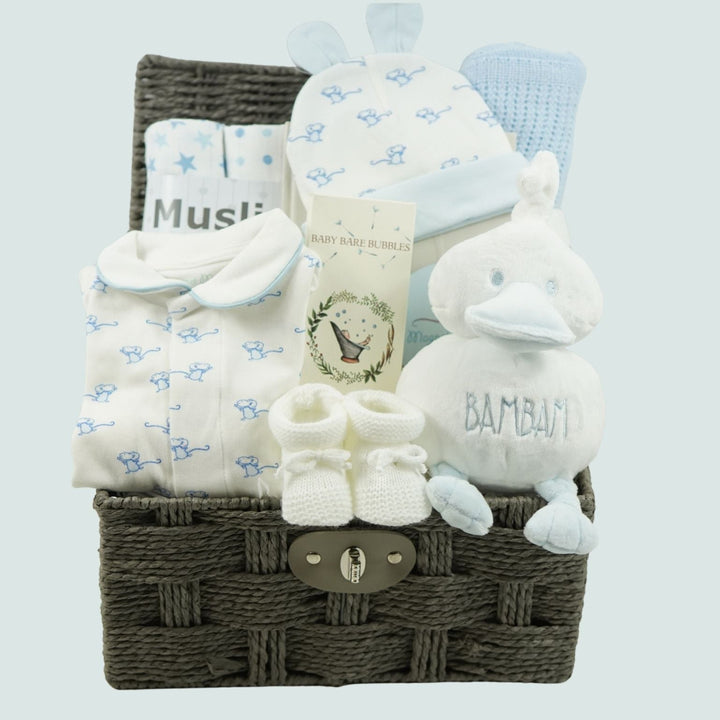 A baby gift basket set containing a white and blue patterned magnetic closing onesie, with mice design and matching hat with ears , a white plush duck toy with 'BAMBAm' embroidered on it, a pair of white knit booties, and a wicker storage basket with a metal clasp, Blue cellular blanket , Muslins And baby toiletries