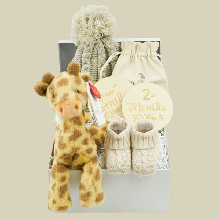 A baby gift set in a white box featuring a plush giraffe toy, cream knit booties, a cream knit pom-pom hat, a cream drawstring pouch, and round tags reading 'smile 2+ months'.