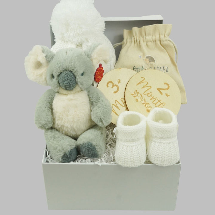 A baby gift set in a white box containing a plush koala toy, a cream-colored knit bootie, a cream drawstring pouch, and two round wooden milestone plaques on a solid gray background.