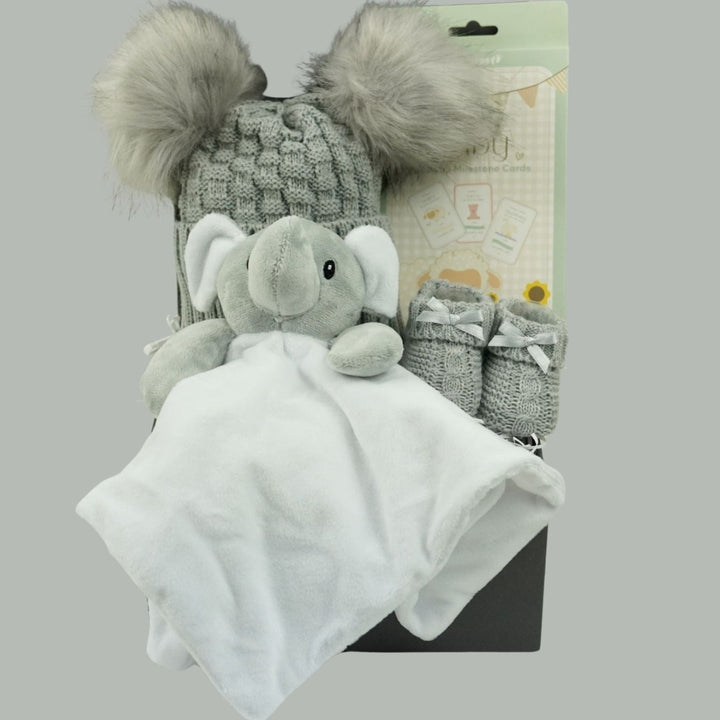 A baby gift set featuring a plush gray elephant comforter, a gray knit beanie with faux fur pom-poms, a pair of gray booties, and a white blanket, displayed against a solid gray background.