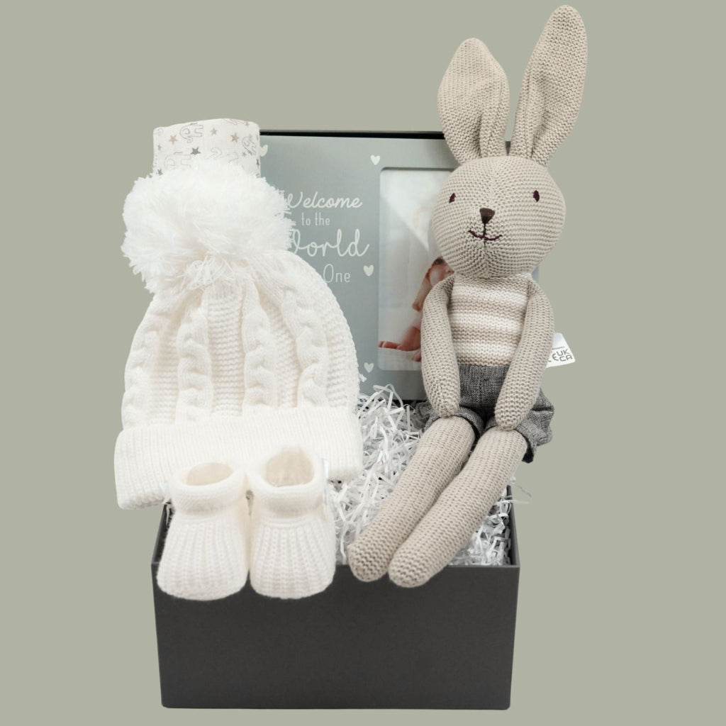 A baby gift set in a grey gift  box featuring a cream knit bonnet with a white pom-pom, matching cream booties, a cream knit hat, and a beige stuffed bunny doll with a striped shirt,  and baby picture frame 