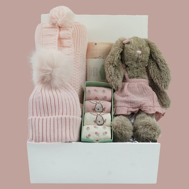 A baby gift basket set in a white box, featuring a pink knit beanie with a fluffy pom-pom, a taupe plush bunny doll in pink overalls, 4 pairs of baby bunny socks  and a folded blanket,