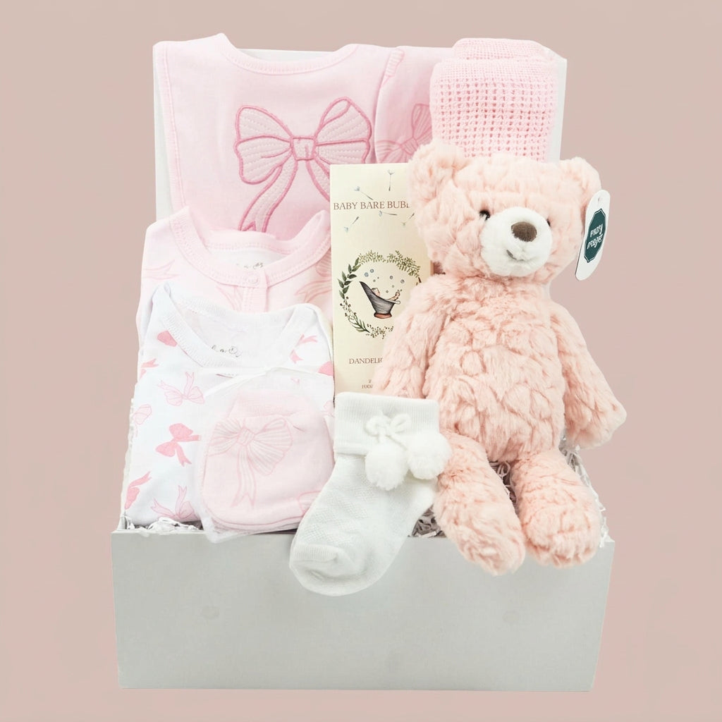 A baby gift basket set in a white box, featuring a light pink long-sleeve bodysuit with a bow, a white bodysuit with pink bows, a pink knit hat, a plush pink teddy bear, a white bootie, and a baby book, set against a solid pale pink background.