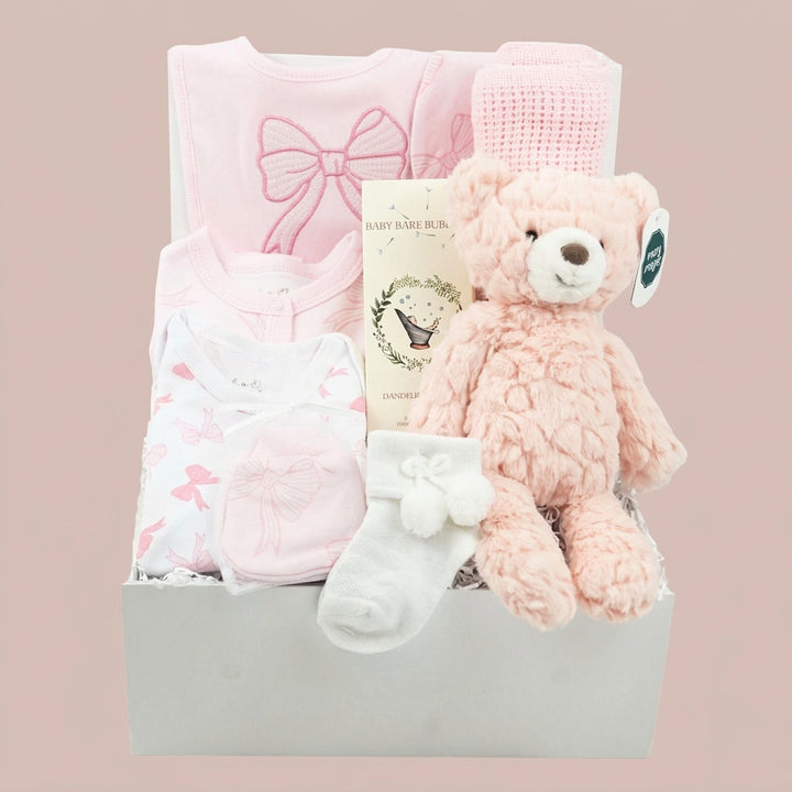 A baby gift basket set in a white box, featuring a light pink long-sleeve bodysuit with a bow, a white bodysuit with pink bows, a pink knit hat, a plush pink teddy bear, a white bootie, and a baby book, set against a solid pale pink background.