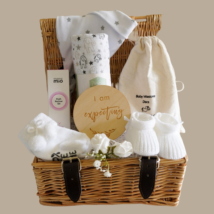 small wicker hamper, perineal oil, wooden pregnancy milestone discs in a cream bag, white with grey elephants baby muslin, white with silver stars baby knot hat, baby socks with cute writing , baby booties 