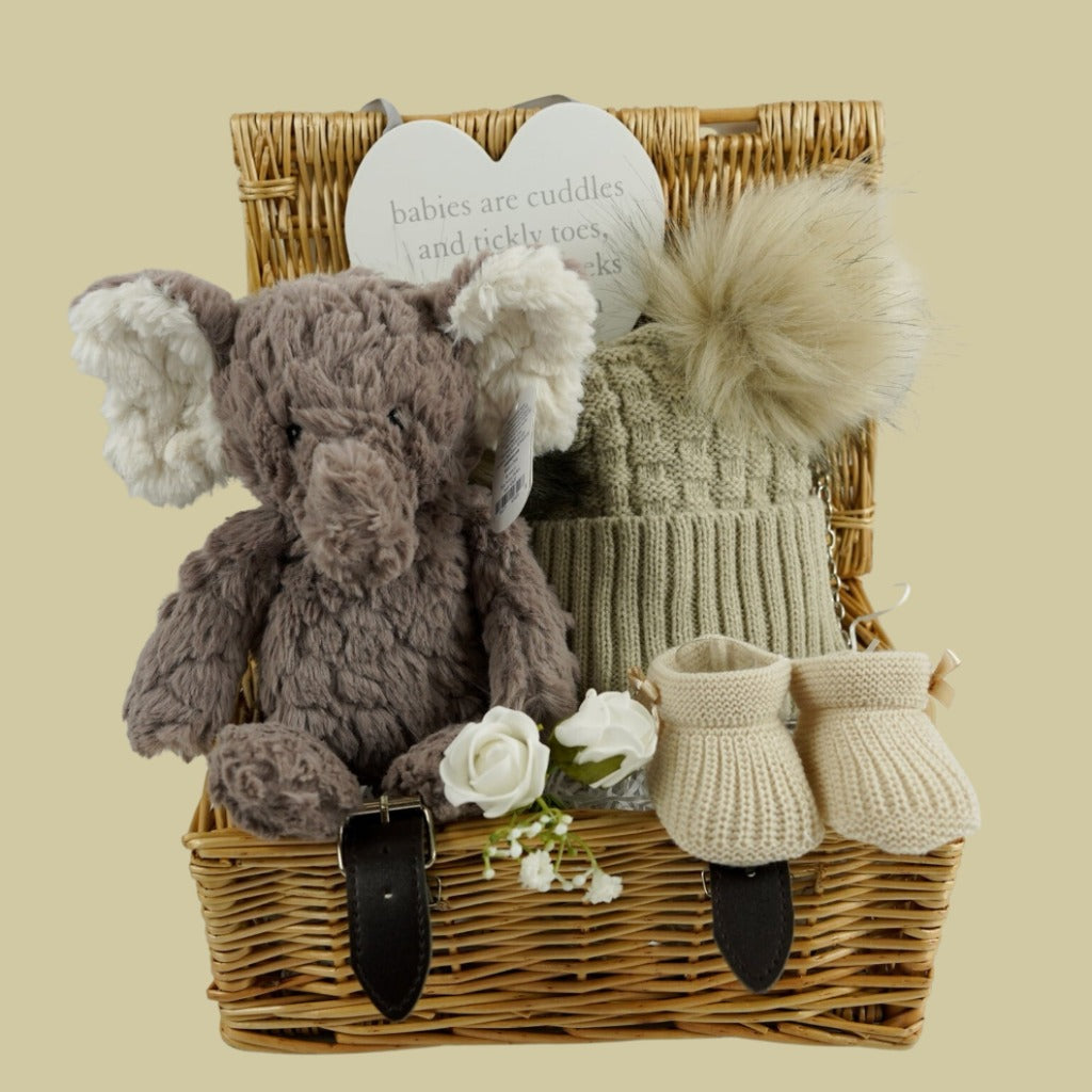 wicker hamper basket with grey elephant soft toy, nursery plaque, biscuit coloured  knit booties, biscuit coloured double fluffy pom pom hat