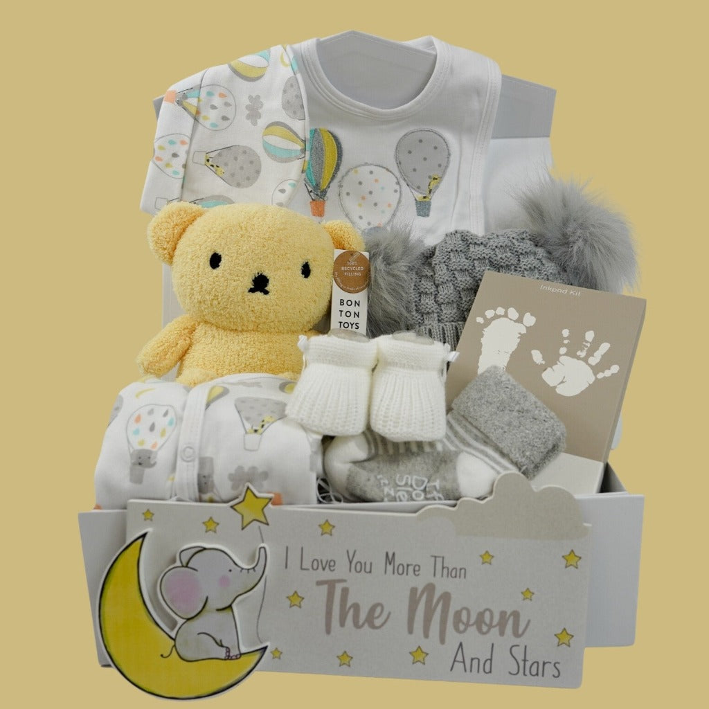 Baby hamper box with yellow Boris bear in terry fabric, white baby sleepsuit with colourful hot air balloons and matching hat, bib, hand and footprint ink pad, baby knit white booties, grey knit hat with double fluffy pom pom